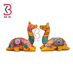 Two hand-painted wooden camel figurines decorated with vibrant pink, green, and orange traditional patterns, set against a plain white background with a KB Art logo in the top-left corner.