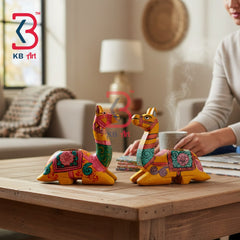 Two hand-painted wooden camel figurines with vibrant multicolor patterns, displayed on a wooden coffee table in a cozy living room setting with a person holding a mug in the background.