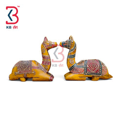 Two hand-painted wooden camel figurines facing each other, decorated with intricate red, black, and white folk art patterns on a yellow-brown base, shown on a white background with a KB Art logo.