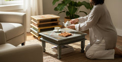 A woman in a light beige robe kneels on a woven rug beside a low wooden coffee table in a bright living room, pouring tea from a white teapot into a cup.