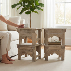 Three carved light gray wooden nesting side tables in a bright living room, with a person seated beside the left table holding a white mug.