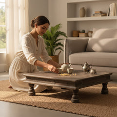 A woman in a cream-colored dress kneels on a woven rug beside a dark wooden coffee table in a bright, modern living room. She is interacting with a small teapot and teacups on the table.