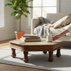 A rustic octagonal wooden coffee table with turned legs in a bright living room. A person's hand is placing a white mug on the table next to a stack of books.