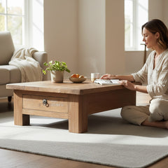 A woman sits on a light gray rug beside a wooden coffee table in a bright living room. The table has one drawer with a metal knob and a small front label that appears to read 'THE TABLE COMPANY.