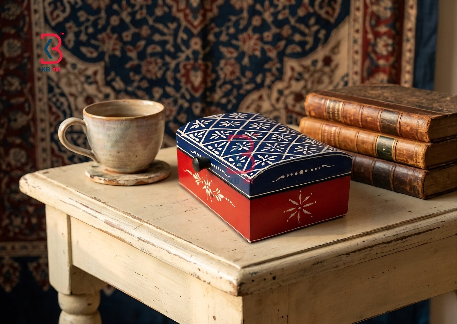 A decorative rectangular metal tin box with a red base and a blue lid featuring white geometric patterns, styled on a distressed white table next to a ceramic cup and a stack of vintage books against a patterned tapestry background.