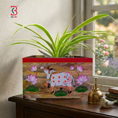 A decorative rectangular planter box with a hand-painted style illustration of a white cow, pink lotus flowers, and green leaves, filled with a green spider plant. The planter sits on a dark wooden table next to a brass sprayer and a small bird figurine, with a window showing a garden view in the background.