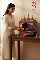 A woman in a cream-colored kurta stands beside a wooden table, offering marigolds to a small, ornate, multicolored Hindu deity home with gold finials. The KB Vision logo is visible in the top right corner.