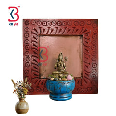 A square carved wooden wall plaque or frame in reddish-brown, styled with a small brass deity statue in a blue pot and a small vase of dried flowers on a white background. KB Art logo is visible in the top-left corner.