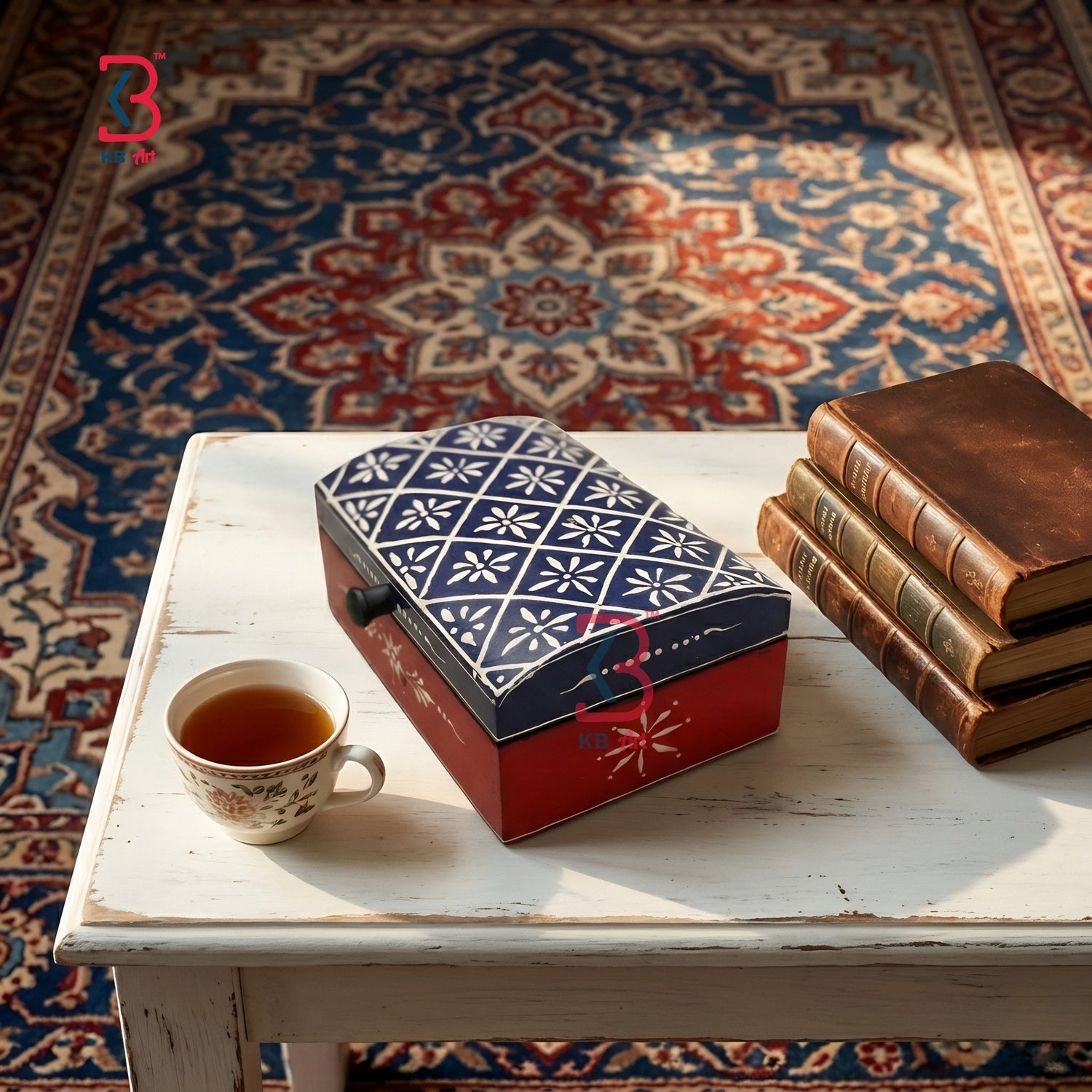 A decorative rectangular tea tin with a blue-and-white floral lid and red base sits on a white table beside a cup of tea and a stack of old books, set on a patterned rug.