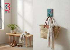 A woven straw tote bag with brown leather handles hangs from a colorful KB Art wall hook in a bright, modern entryway. The right side shows the hook mounted on a white wall with the bag hanging, and the left side shows a styled entryway with a wooden bench, plant, and boots.