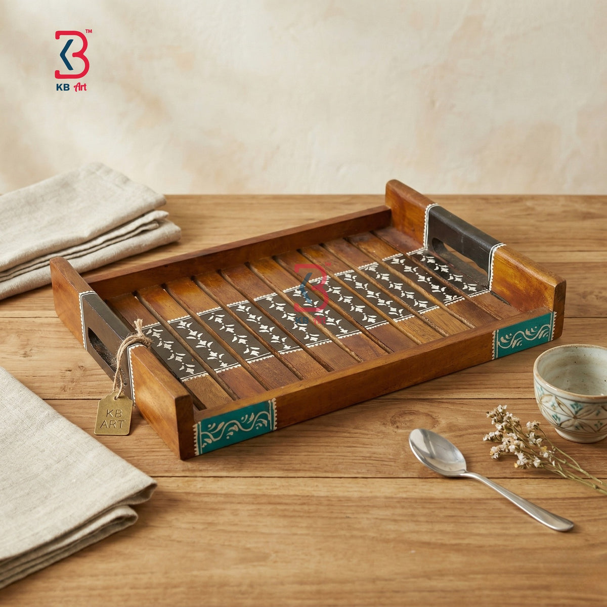 A rectangular wooden serving tray with decorative inlay details, shown on a wooden tabletop with folded napkins, a small bowl, and a spoon nearby. A KB retail logo appears in the top left corner.