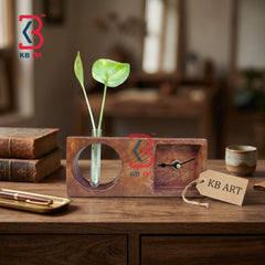 A rustic wooden desk clock and vase set on a wooden table, featuring a rectangular wood block with a circular cutout holding a green plant stem and a small square compartment with a clock face. A KB Art branded tag hangs from the front, with books and a cup in a softly blurred background.
