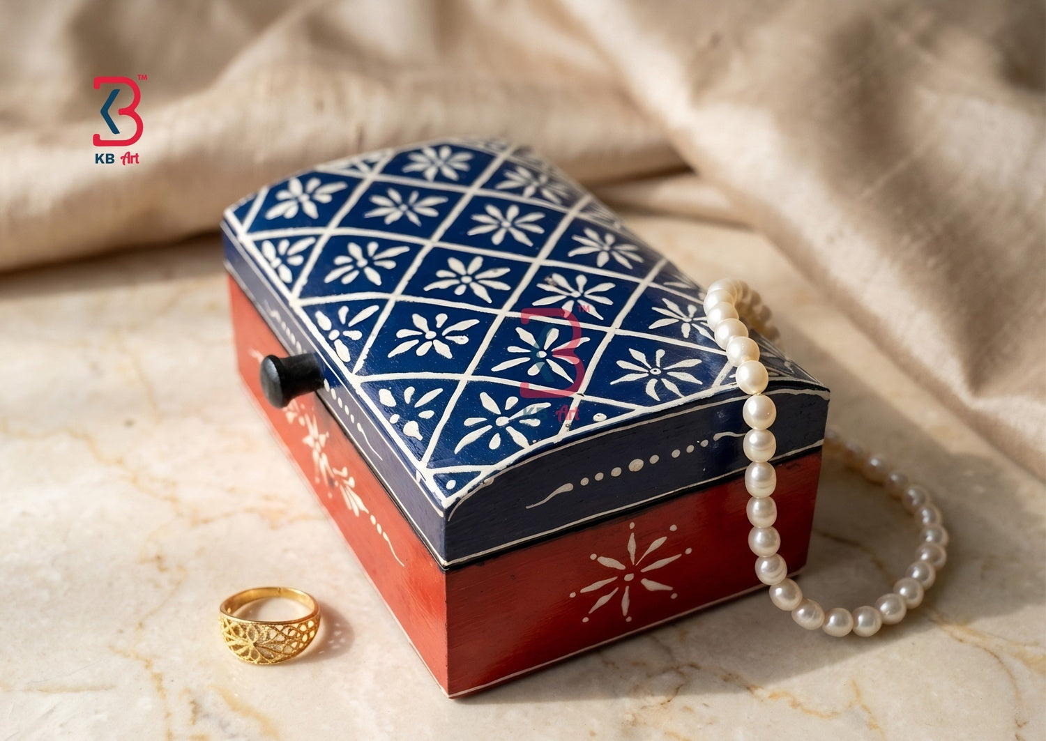 A decorative jewelry box with a navy blue and white floral patterned lid and a red base, styled on a beige marble surface with a pearl strand draped over it and a gold ring nearby.