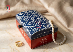 A decorative jewelry box with a navy blue and white floral patterned lid and a red base, styled on a beige marble surface with a pearl strand draped over it and a gold ring nearby.