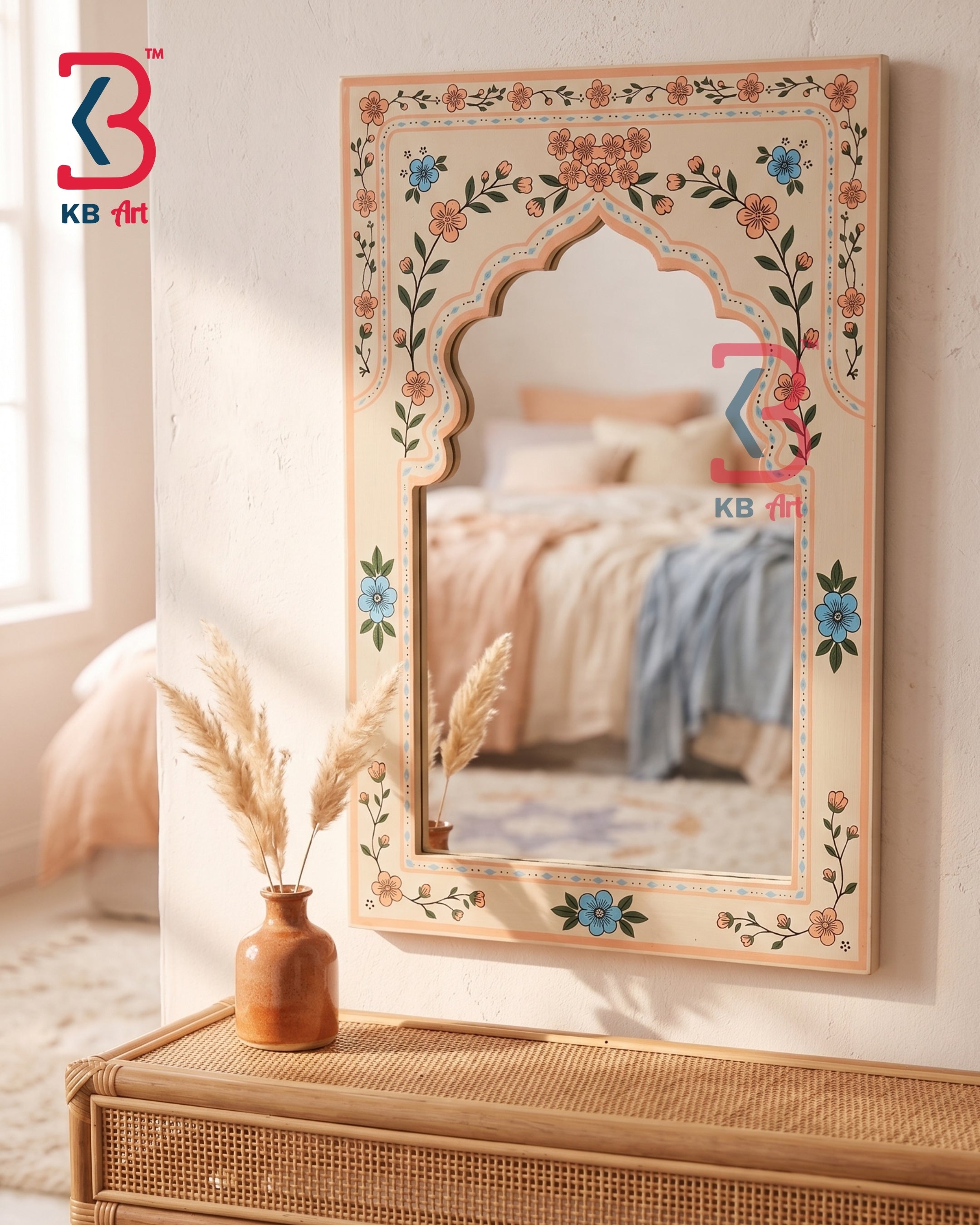 An ornate arched wall mirror with a beige frame decorated with colorful floral and vine motifs, hanging on a textured wall above a woven rattan console table. The mirror reflects a softly lit bedroom with pink and blue bedding, and a small vase with dried grass sits on the table.
