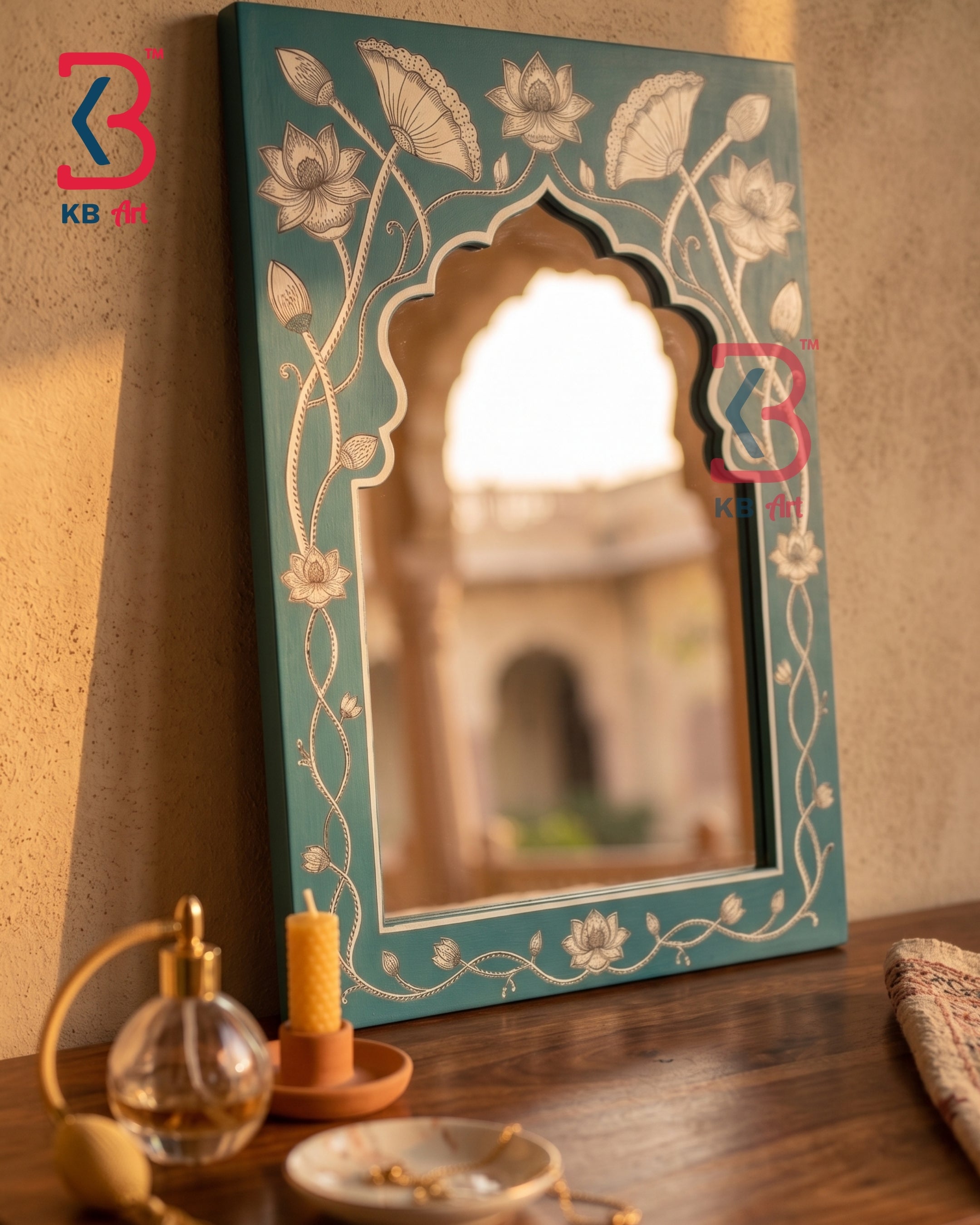 A decorative arched wall mirror with a teal painted frame and white floral vine motifs, leaning on a wooden surface against a warm beige wall. The mirror reflects a softly blurred courtyard scene, and small tabletop decor items are visible in the foreground.