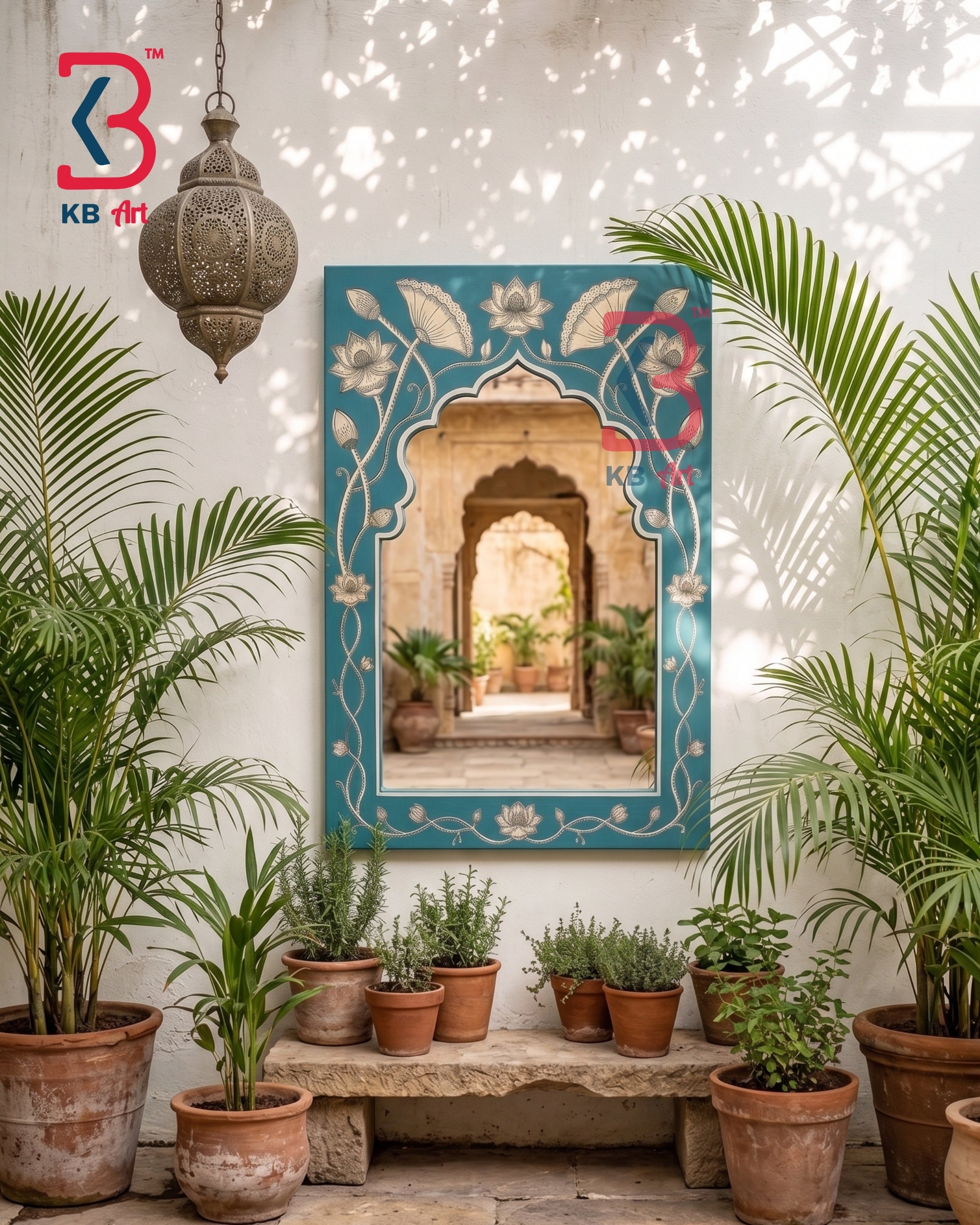 A decorative wall mirror with an ornate turquoise blue border featuring white floral and vine motifs, mounted on a white stucco wall. The mirror reflects an arched corridor with plants, and the scene is styled with potted palm trees, small potted herbs on a stone bench, and a hanging lantern.