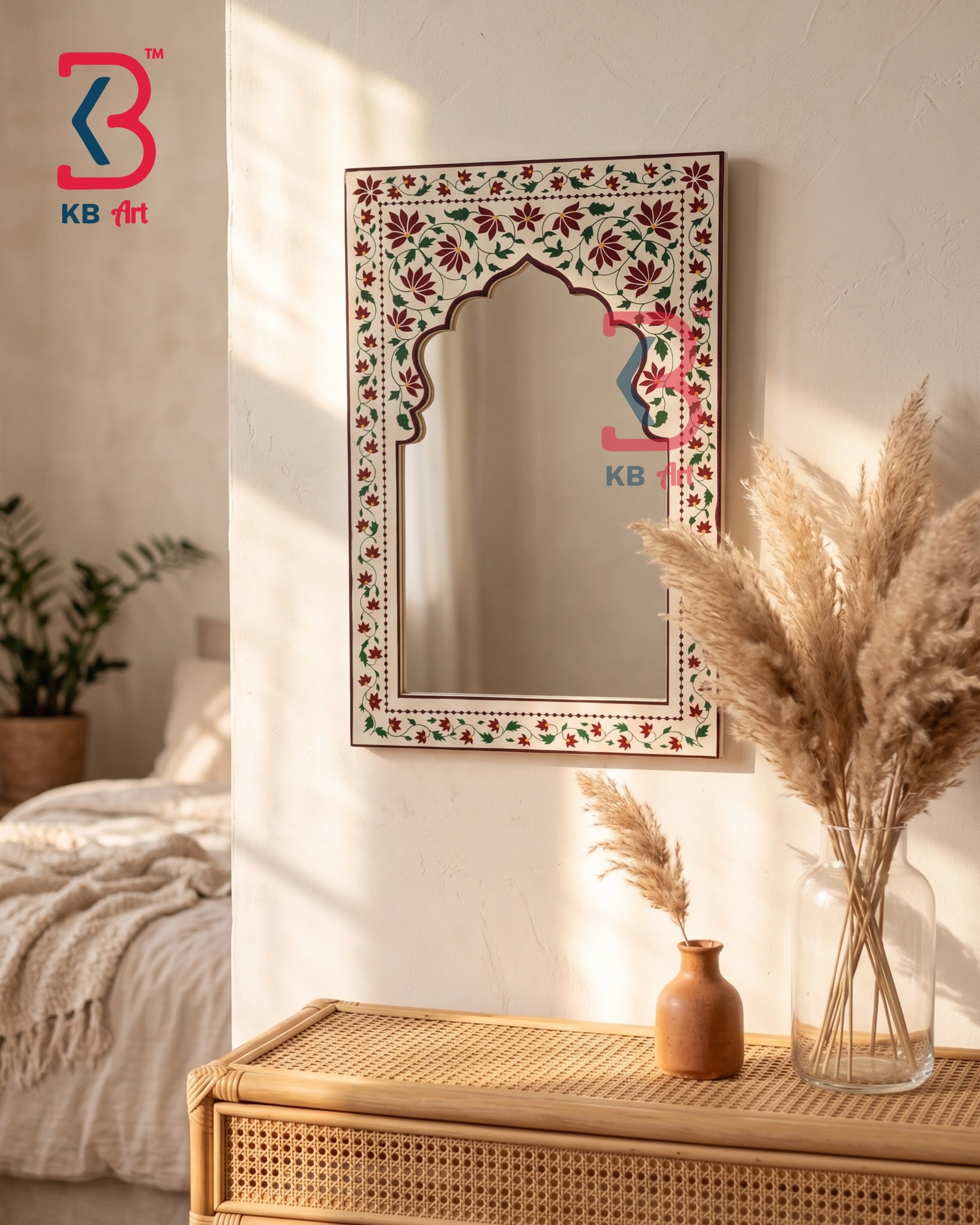 An ornate rectangular wall mirror with a floral hand-painted frame hangs on a textured cream wall above a rattan console table. The scene is styled with a glass vase of pampas grass and a small terracotta pot, with soft sunlight casting shadows across the room.