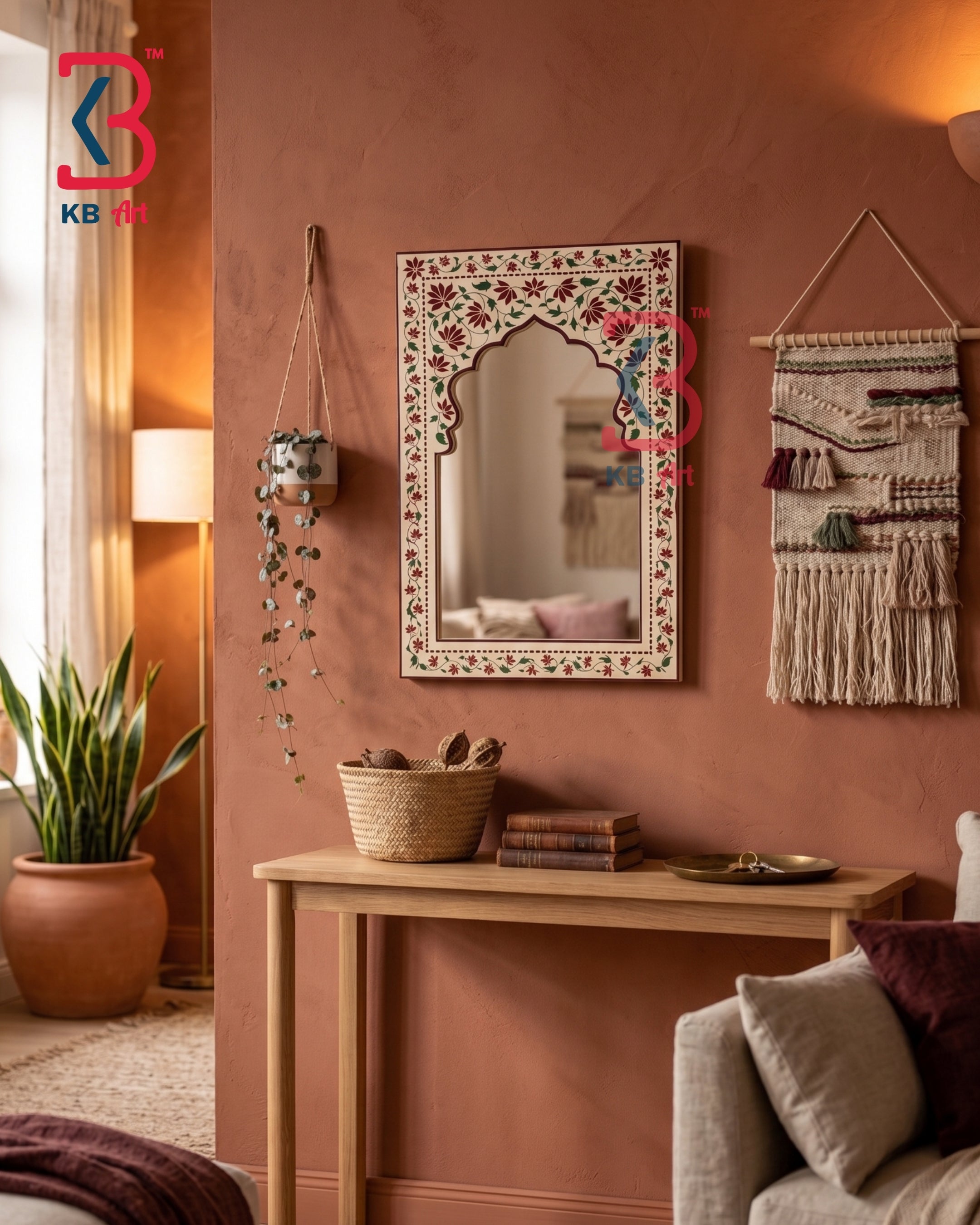 An ornate rectangular wall mirror with a floral bohemian-style frame hangs on a terracotta-colored wall in a styled living room setting. A light wood console table, a potted snake plant, a woven basket, and a gray sofa are visible in the room.