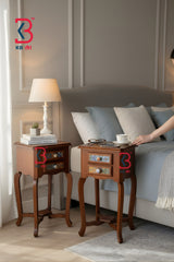 A bedroom scene showing two wooden nightstands beside a bed. One nightstand holds a white table lamp and books, while the other has a small tray and a hand reaching toward it.