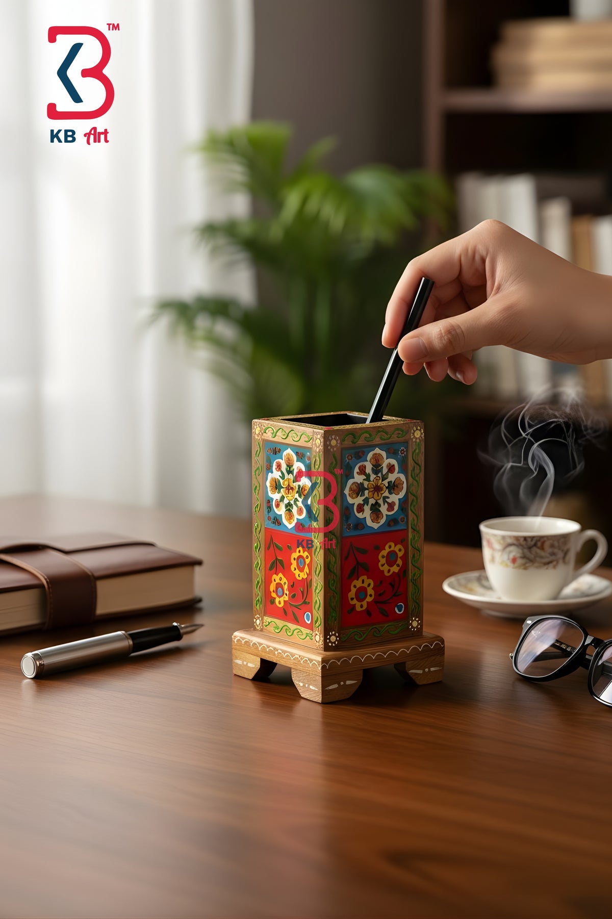A hand inserts a black pen into a decorative wooden pen holder with colorful floral patterns, set on a wooden desk with a notebook, glasses, and a cup of tea in a warm, blurred office environment.