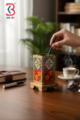 A hand inserts a black pen into a decorative wooden pen holder with colorful floral patterns, set on a wooden desk with a notebook, glasses, and a cup of tea in a warm, blurred office environment.