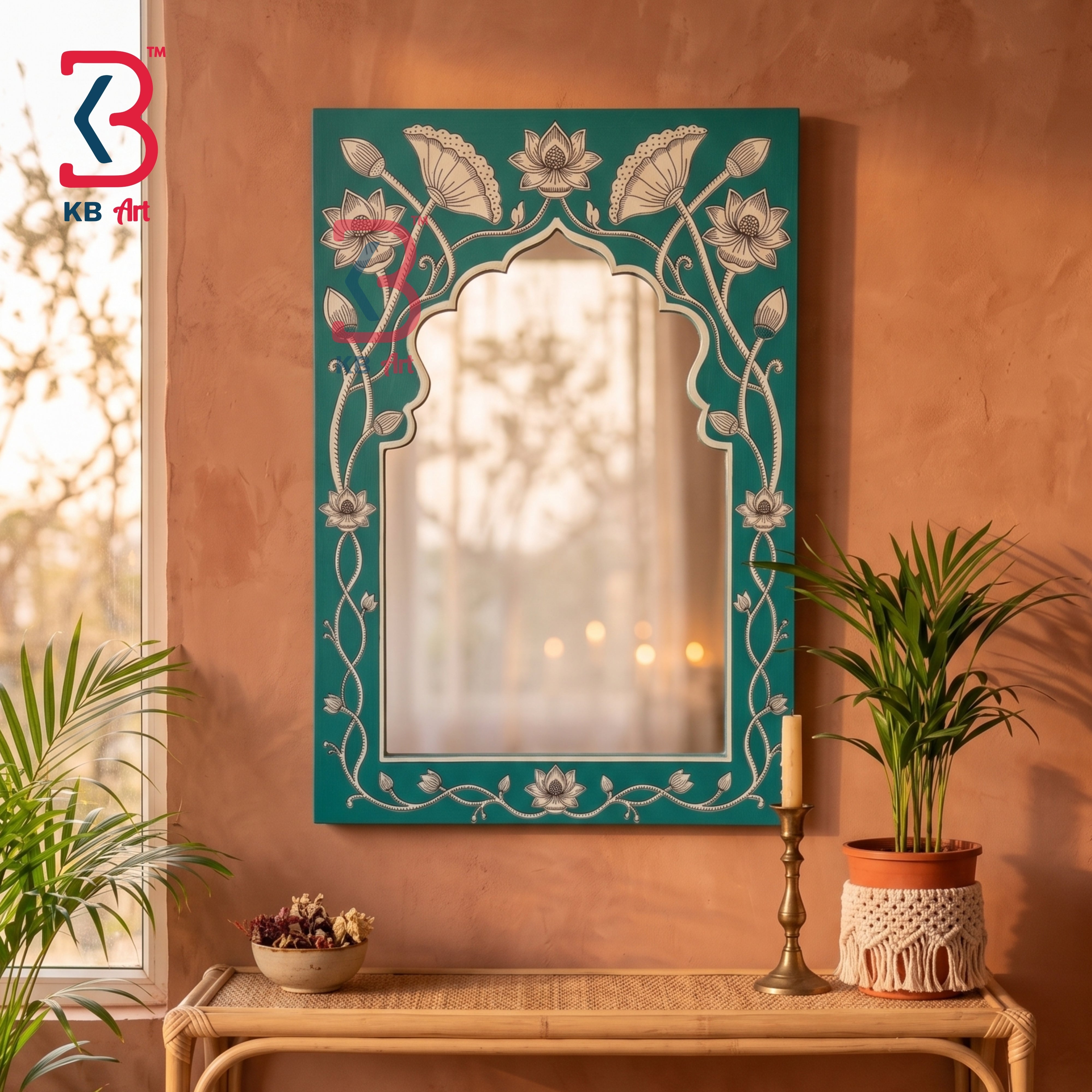 A decorative arched wall mirror with a teal border and white floral vine inlay, displayed on a terracotta wall above a woven console table with potted plants and a candle.
