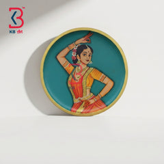 A round decorative plate featuring a painted traditional Indian dancer in a red and orange sari against a teal background, with a gold rim, photographed on a white surface with a cast shadow.