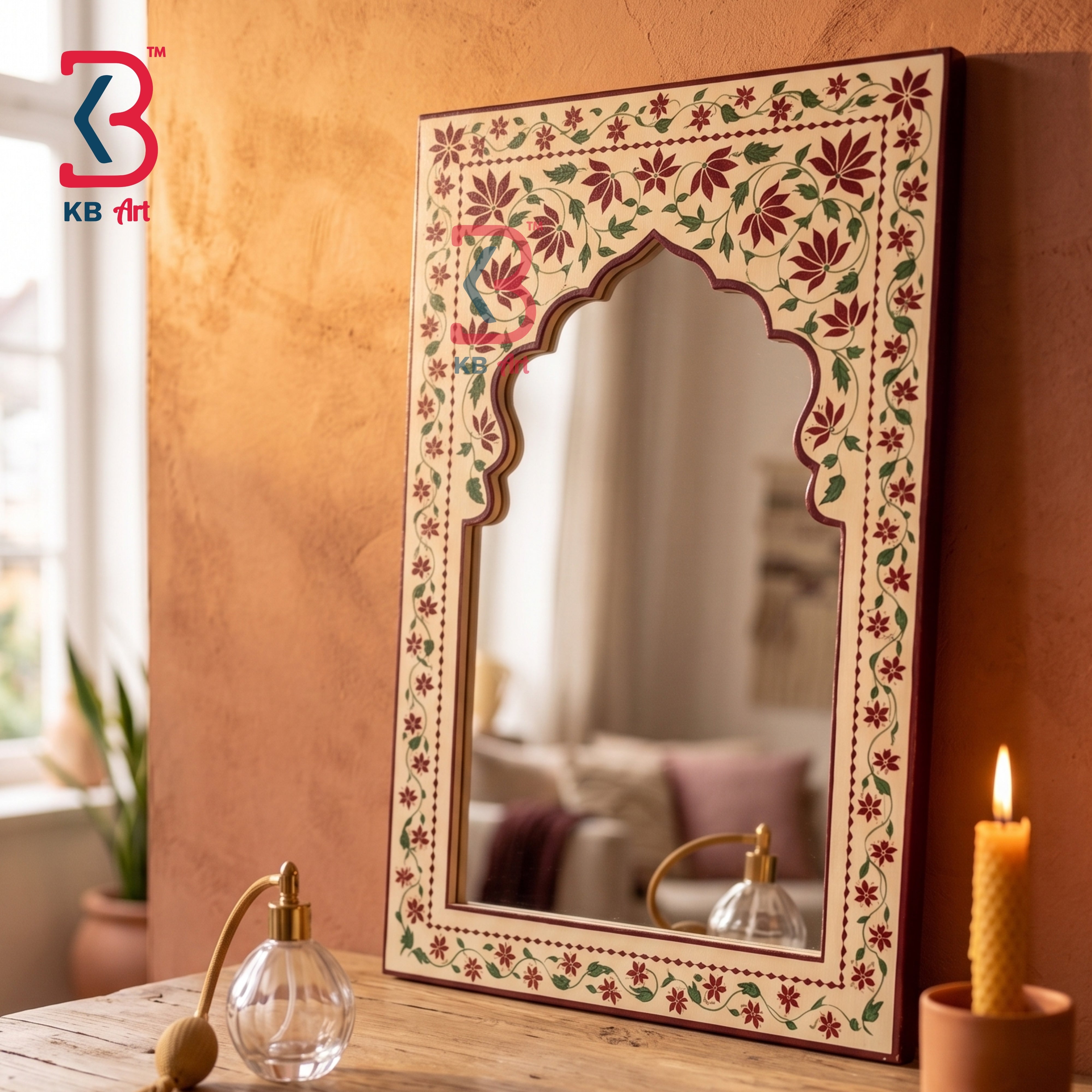 A decorative rectangular wall mirror with an ornate arched top and a hand-painted floral border, displayed on a wooden table in a warmly lit room. The mirror reflects a softly blurred interior, and nearby props include a glass perfume bottle, a lit candle, and a small plant.