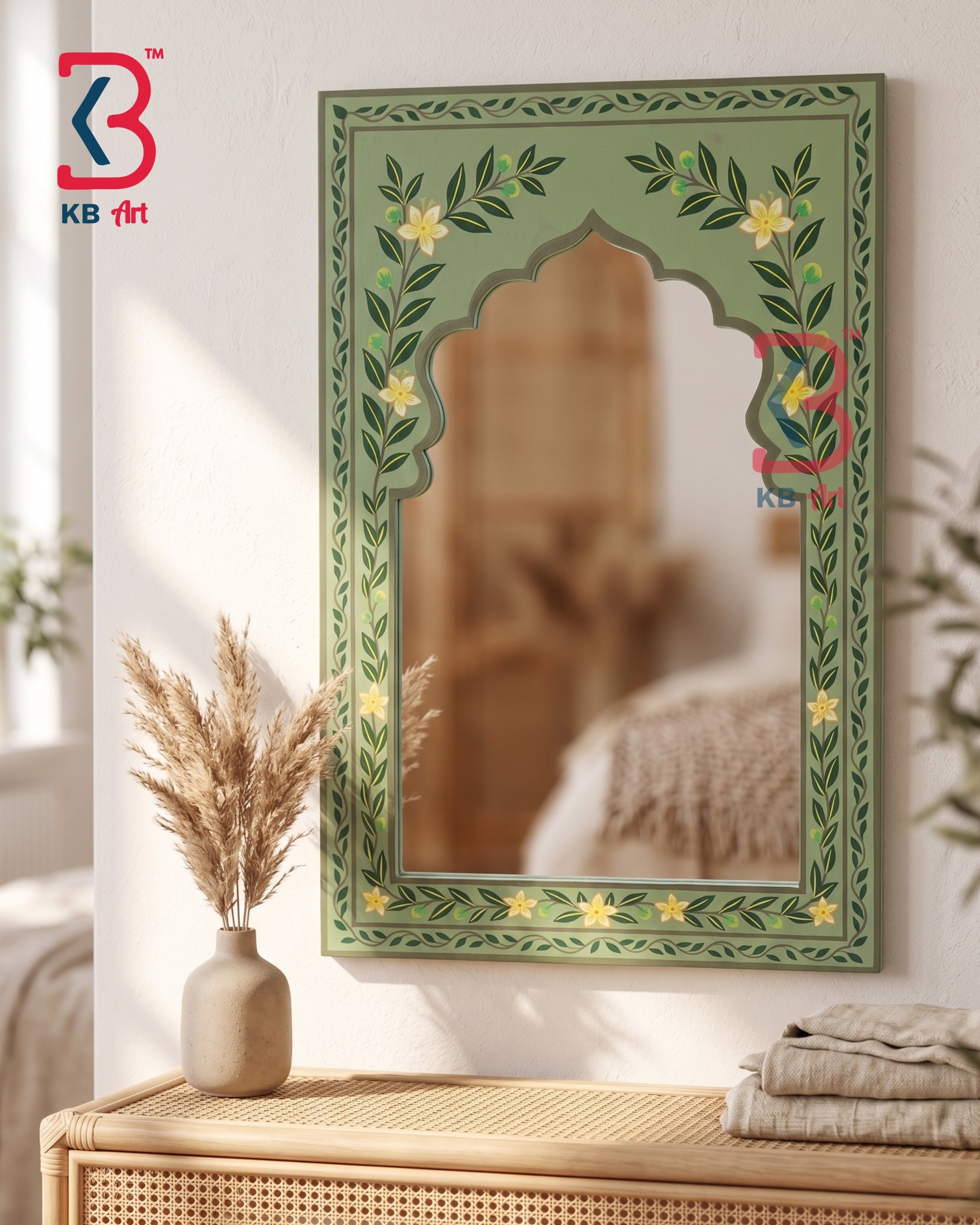 An ornate green arched decorative wall mirror with floral and vine border details, shown hanging on a light wall above a woven console table with dried grass in a vase and folded linens. A KB art watermark is visible in the top-left corner.