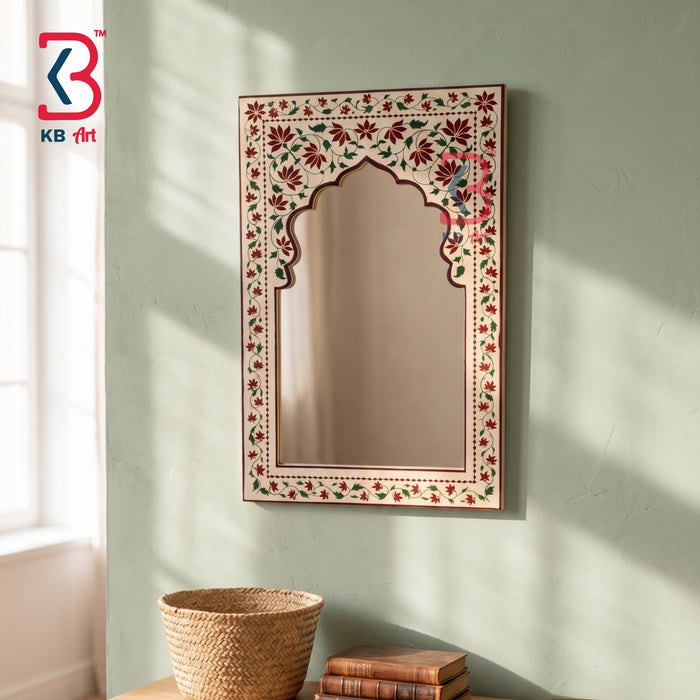 An ornate rectangular wall mirror with an arched center and a hand-painted floral border, mounted on a textured sage green wall. The mirror is styled in a room setting above a wooden table with a woven basket and books, with soft sunlight casting shadows on the wall.