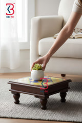 A dark brown wooden low coffee table with a colorful mosaic tile top, shown in a living room setting with a person's hand placing a small potted succulent on it.