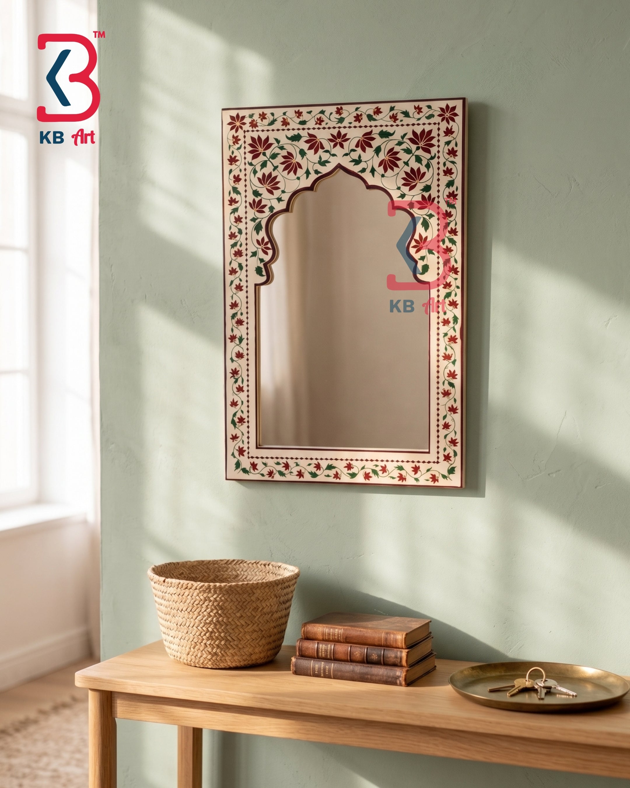 An ornate rectangular wall mirror with a floral arch design in a cream, red, and green hand-painted style, hanging on a sage green wall above a wooden console table with a woven basket and books.