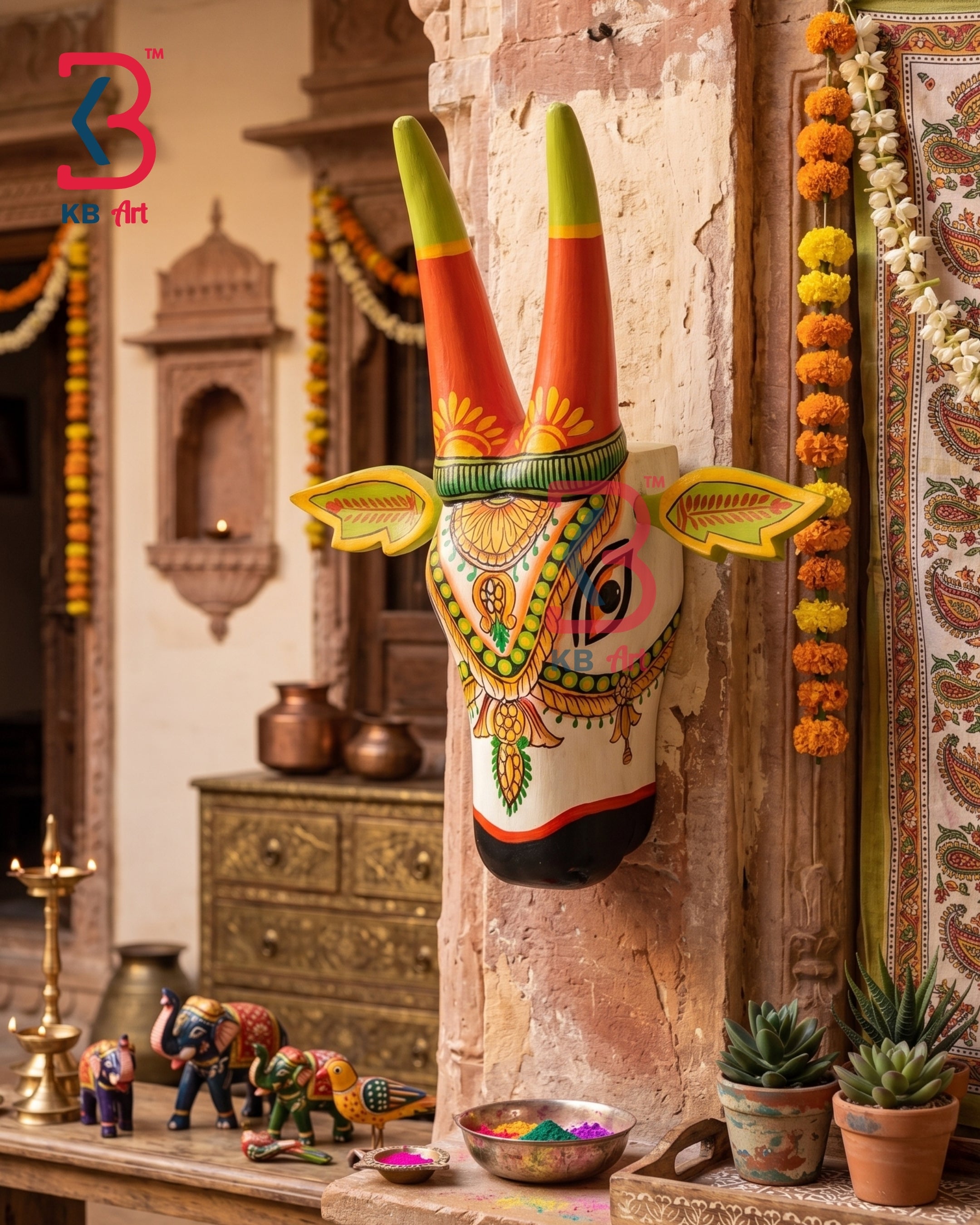 A hand-painted traditional Indian Golu wall hanging depicting a stylized bull head with long orange and green horns, mounted on a rustic stone pillar in a decorated home interior.