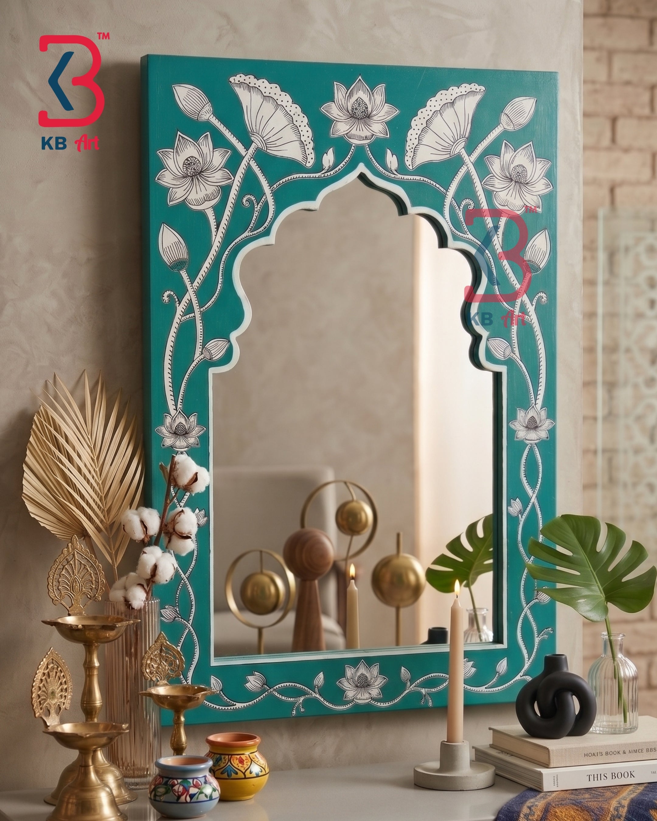 A decorative arched mirror with a turquoise frame featuring white floral and vine motifs, styled on a tabletop with candles, books, and plants in an indoor setting.