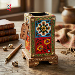 Hand-painted wooden pen holder with traditional Indian floral motifs, displayed on a rustic wooden table with books, a fountain pen, dice, and a cloth in the background.