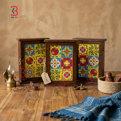 Three small wooden decorative boxes with colorful mosaic tile-style front panels are arranged on a rustic wooden table. The scene includes a brass oil lamp, a woven basket with walnuts, pinecones, and a blue fringed cloth, against a warm beige wall.