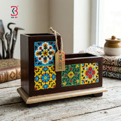 A dark brown wooden desk organizer with colorful hand-painted floral tile panels on the front, resting on a rustic wooden table. A small hanging tag reads 'KB ART'.