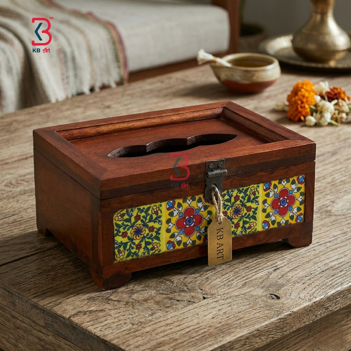 A rectangular wooden tissue box holder with a carved slot on top, decorated with colorful floral mosaic panels on the front. It sits on a rustic wooden table in a styled indoor setting with a bowl, flowers, and a blanket in the background.