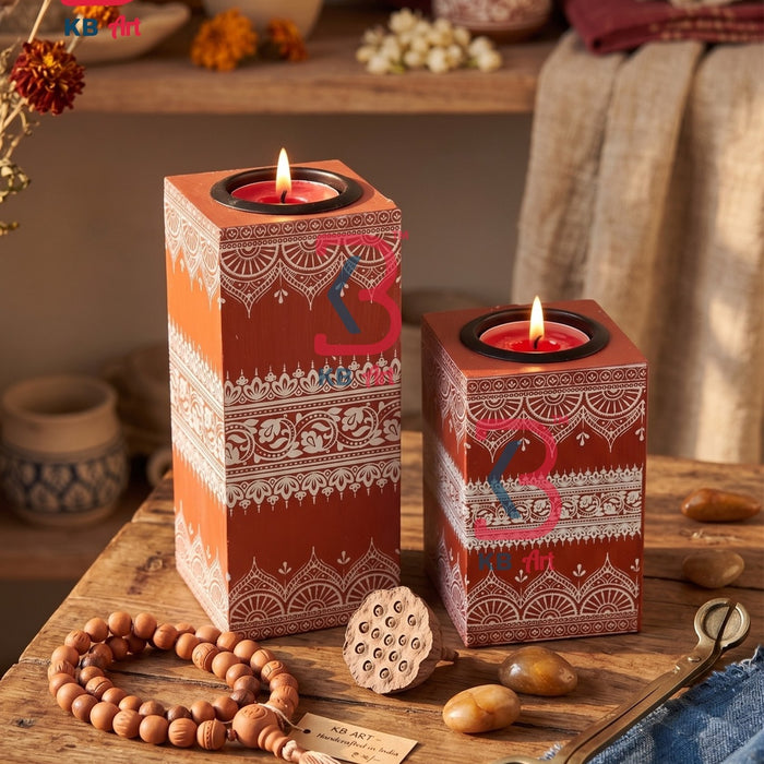 Two decorative rectangular candle holders with red and orange geometric patterns, holding lit tealight candles, displayed on a rustic wooden table with a beaded necklace, scissors, and dried flowers in a warm, bohemian setting.