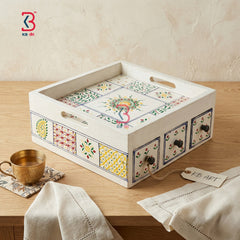 A decorative rectangular serving tray with cutout handles and small front drawers, decorated with colorful geometric and floral tile-style patterns on a light wood table. A brass cup, folded cream cloth, and a KB Art tag are also visible in the styled setting.
