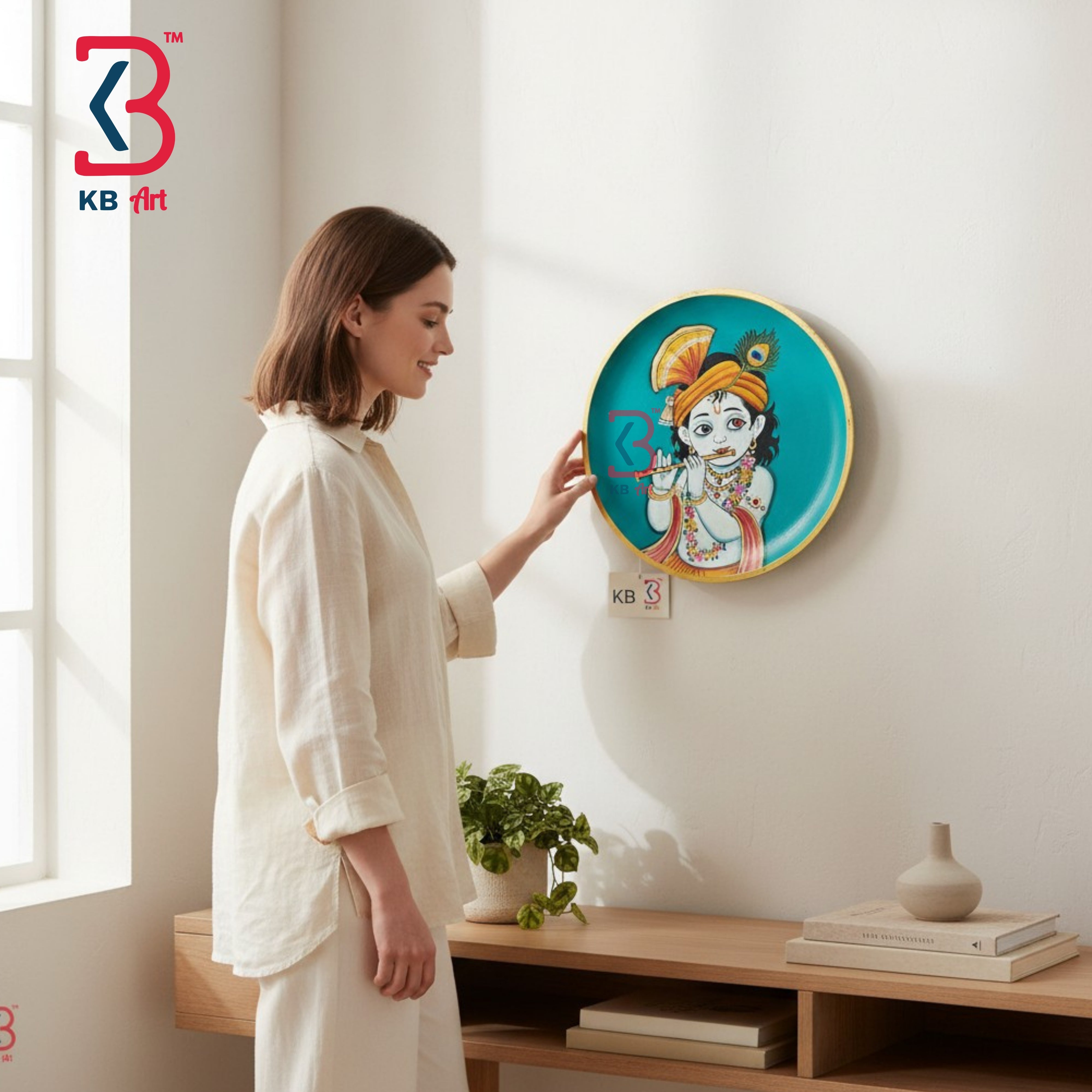 A woman in a cream linen outfit stands in a bright room, touching a round turquoise wall art piece featuring a stylized child character. The artwork has a gold border and is mounted on a white wall above a wooden shelf with books and a small plant.